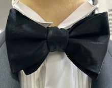 Load image into Gallery viewer, A close-up image of a black butterfly bow tie tied around a white shirt collar, with a person wearing a suit in the background.